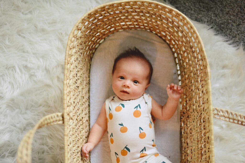A baby wearing a pineapple onesie is lying in a wicker moses basket